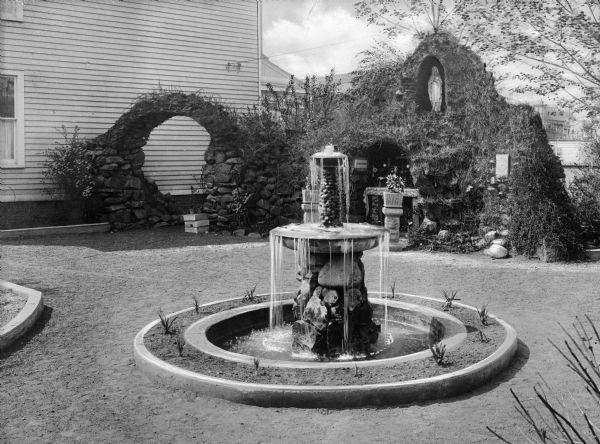 A view of the stone fountain and grotto at Our Lady of Wisdom Academy. The grotto contains an altar to the Virgin and an alcove at the top contains a statue. A clapboard-covered building is on the left.