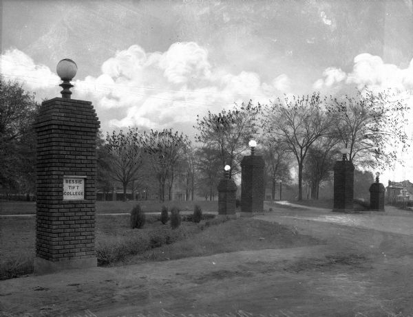 A view of the Carithers Memorial Gate, at the entrance to the Bessie Tift College campus, consisting of brick pillars with globe lamps at the top. The pillar in the left foreground has a sign that reads: "Bessie Tift College." Several campus buildings are in the background.