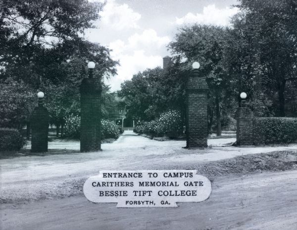 A view of the Carithers Memorial Gate at the entrance to the Bessie Tift College campus, consisting of four brick pillars with globe lamps on top flanking the main pathway into campus. Trees and shrubs line the walkway, with a campus building in the background. A plaque printed in the middle reads: "Entrance to Campus Carithers Memorial Gate Bessie Tift College, Forsyth, GA."