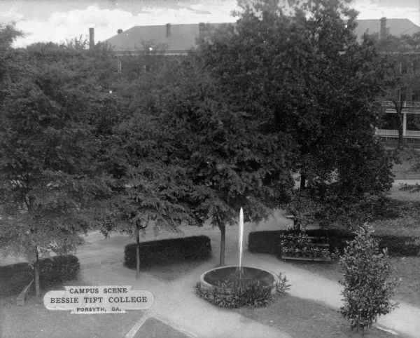 A view of an outdoor campus area taken from an elevated vantage point. Various paths and a row of hedges surrounds an area containing trees, benches, plantings, and a fountain. A college building is partially visible in the background. Printed in the bottom left corner: "Campus Scene Bessie Tift College Forsyth, GA."