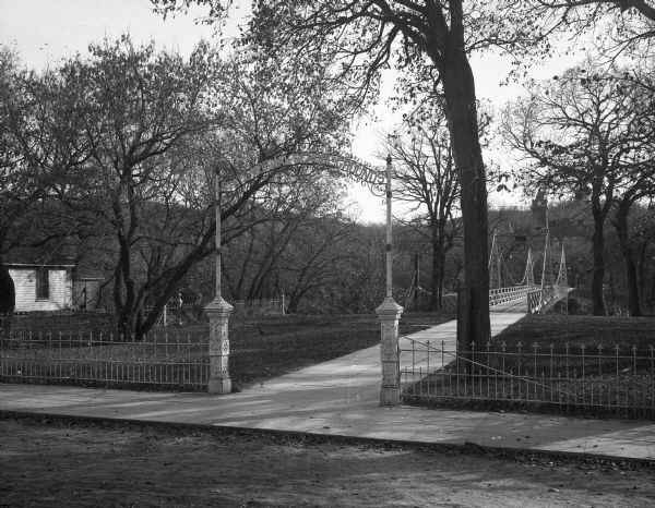 A view of the entrance and foot bridge to the campus at State Normal School. A wrought iron gate surrounds the property, and a metalwork banner over the entrance reads: "State Normal School Grounds." Trees dot the landscape, and a wooden house is to the left, along with a college building in the background. A sidewalk leads to the entrance.