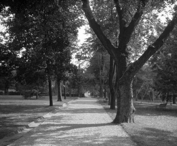 View of a tree-lined walkway to Mother Seton's Spring at St. Joseph's College. A swing set and bench are on the right, and a line of shrubs is in the distance.
