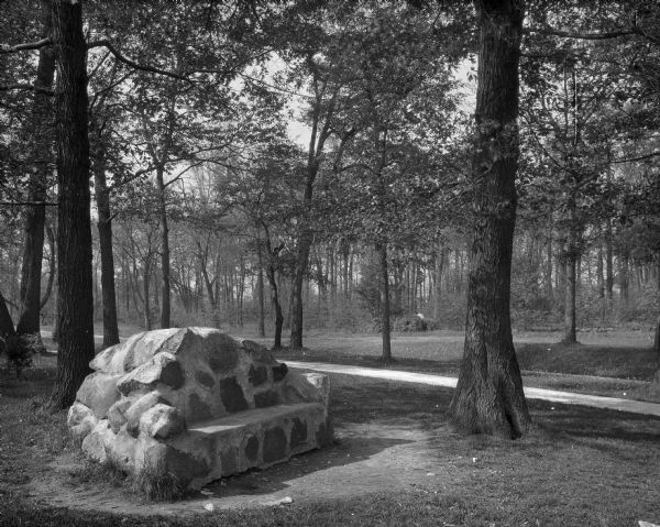 A view of the Freshman Seat, consisting of stones mortared together, situated in a wooded area near a path at Northern Illinois State Normal School.