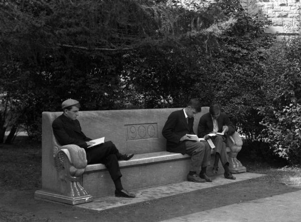 A view of three men sitting outdoors and reading on the Senior Bench at the University of Illinois. The seat is made of stone and "1900" is engraved into its back.