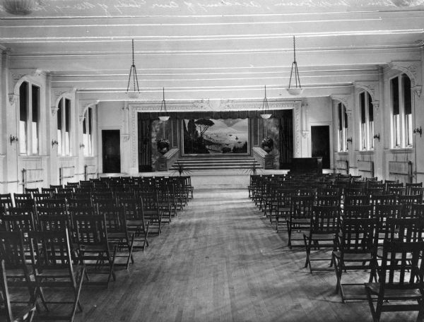 View down center aisle in the auditorium at the Academy of Our Lady of Mercy toward the stage. The audience chairs are arranged in rows. Windows line both the left and right sides of the space and a piano stands to the right side of the stage. The stage set depicts stone steps that overlook a body of water and mountains.