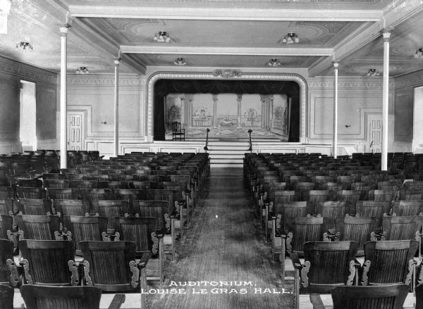 View down central aisle in the auditorium at the College of Mt. St. Vincent toward the stage. A central set of steps leads up to the stage. The stage is set with a backdrop depicting an interior looking out onto a garden. Caption reads: "Auditorium. Louise Le Gras Hall."