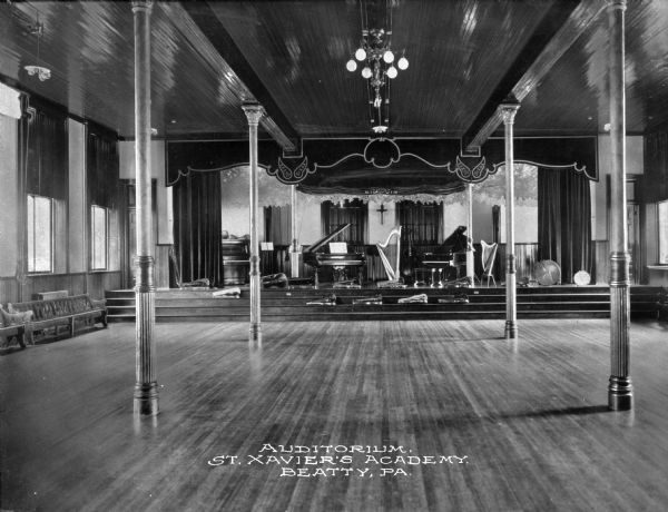 A view of the auditorium at St. Xavier's Academy. Wooden benches line the walls and both the left and right walls contain windows. The stage appears to be set for a musical performance, complete with pianos, harps, violins, and cellos. Caption reads: "Auditorium. St. Xavier's Academy. Beatty, PA."