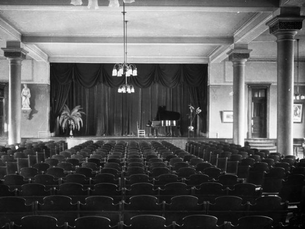 A view of the auditorium at Seton Hill College, looking towards the stage from behind rows of wooden seats. The stage is set with a piano and potted plants.