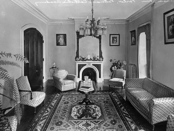 A view of the parlor at Putnam Catholic Academy. A small table stands on a carpet in the middle of the room, surrounded by upholstered chairs and a sofa. The fireplace mantle contains small decorative objects and the walls are decorated with a mirror and several paintings.