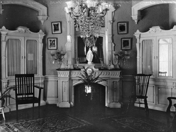 A view of the parlor's fireplace and mantle, with cabinets standing on either side. A statue of the Virgin sits in the mantle along with candlesticks. Decorative light fixtures flank the sides of the mirror above the mantle.