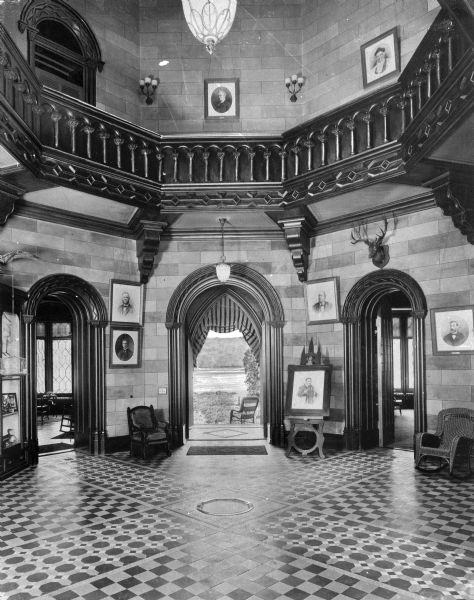 A view of the rotunda at Fonthill Castle, with the floors decorated with parquetry. Two doorways lead to additional rooms while another in the center is open to the outdoors. The rotunda is decorated with many prints and objects, including a mounted deer head.