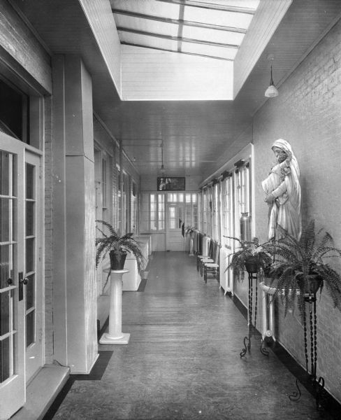 A view of the classroom hallway at Our Lady of Wisdom Academy,  brightly lit by large windows and skylights. Potted plants on stands line the hallway, and a statue of Mary holding Jesus is the main decoration.