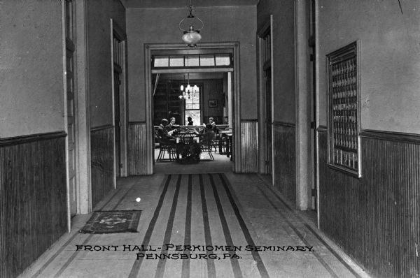 View down the front hall at Perkiomen Seminary, towards a room with a group of men reading around a table. Hall furnishings include two carpets and a group of framed photographs. Caption reads: "Front Hall-Perkiomen Seminary. Pennsburg, PA."