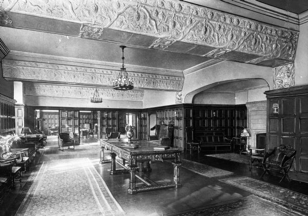 View of the lounge in Ida Nayos Hall at the University of Chicago.  A large tables sits in the middle of the room and upholstered chairs are arranged near bookcases lining the walls. An alcove to the right houses a fireplace, and decorative molding lines sections of the ceiling.