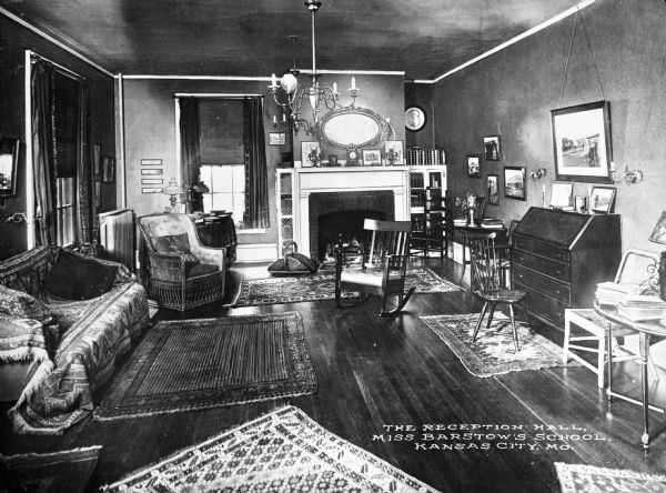 A view of the reception room at Miss Barstow's School. Bookcases with glass fronts flank the fireplace and upholstered and wooden chairs are arranged throughout the room. The mantle contains a clock and framed illustrations, and a mirror hangs above it. Caption reads: "The Reception Hall, Miss Barstow's School. Kansas City, MO."