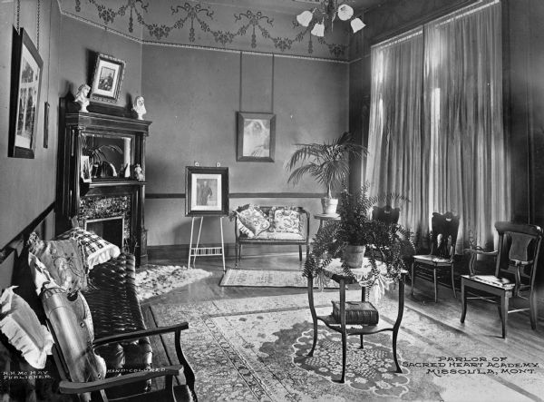 A view of the parlor at Sacred Heart Academy. The room is furnished with a small table which holds a potted plant and Bible. Lining the walls are upholstered and non-upholstered seating. The fireplace is tiled, and the mantle contains a print and small figurines. Prints also decorate the walls. Caption reads: "Parlor of Sacred Heart Academy. Missoula, Mont."