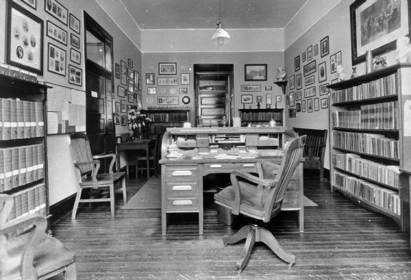 A view of the headmaster's office at Pennington Seminary. Bookcases and chairs line the walls, and a desk sits in the middle of the room. Framed photographs and prints decorate the walls.
