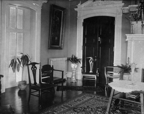 View of the Bishop's parlor at the Academy of Our Lady of Mercy. A table with vase sits near the fireplace and has chairs gathered near it. A potted plant sits near the shuttered window and a painting hangs on the wall above a radiator.