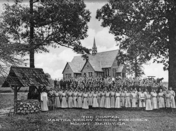 Students and staff pose for a group portrait in front of a wooden chapel. A well stands to their left. Caption reads: "The Chapel, Martha Berry School for Girls.  Mount Berry, GA."