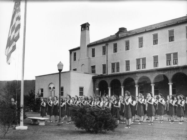 Female students recite the Pledge of Allegiance while standing in formation near a flag pole outside Immaculate Heart Academy. The school building, with an arched column porch, is behind the girls.