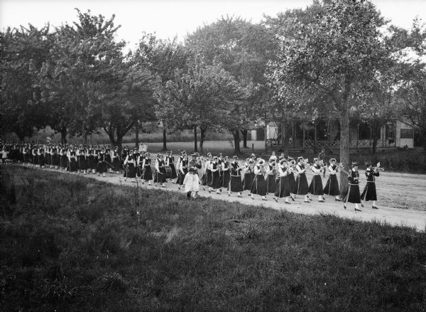 Elevated view of the women's marching band from John Wanamaker Commercial Institute performing, with children walking beside them and standing on the side of the road. The women wear uniforms with long skirts and vests.