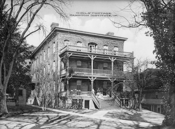 A view of the female residence at Hampton Institute. The left side of the front porch contains potted plant,s and three women converse near the steps. Another building is on the right. Caption reads: "Girls' Cottage, Hampton Institute, VA."