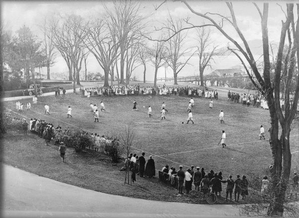 Elevated view of women wearing uniforms of knee-length skirts and dark tights taking to the field to play a game of field hockey at Hampton Institute. Spectators have gathered on all sides to watch. In the background is a dock and lake.