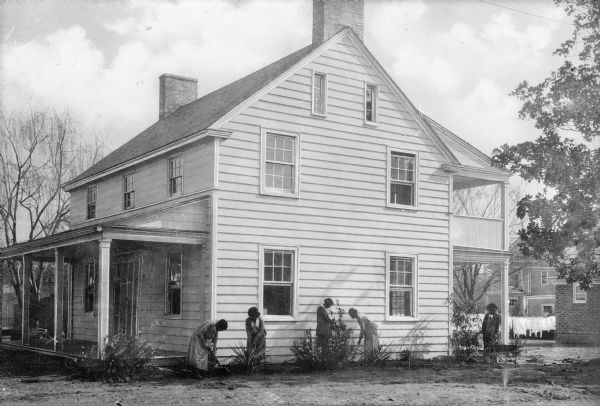 Female students garden next to the Home Economics Practice House. The house has clapboard siding on its two stories and there is a porch on the front and back. The home represents the "Farmhouse Style," made specifically to address rural housing needs. Garments dry on a clothing line behind the house.