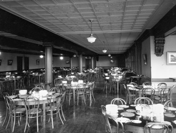 A view of the dining room at State Teacher's College, with rows of round tables lined across the space. Each table is set with a placemat and silverware setting for each chair. Bowls and plates are stacked in the middle of each table.