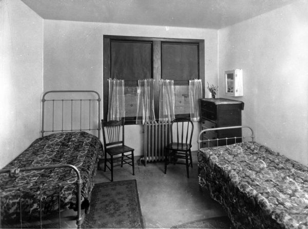 A view of a bedroom at Switzer Foundation for Girls furnished with two twin beds, two rugs, two chairs, and a bureau. The back wall has a large double window.