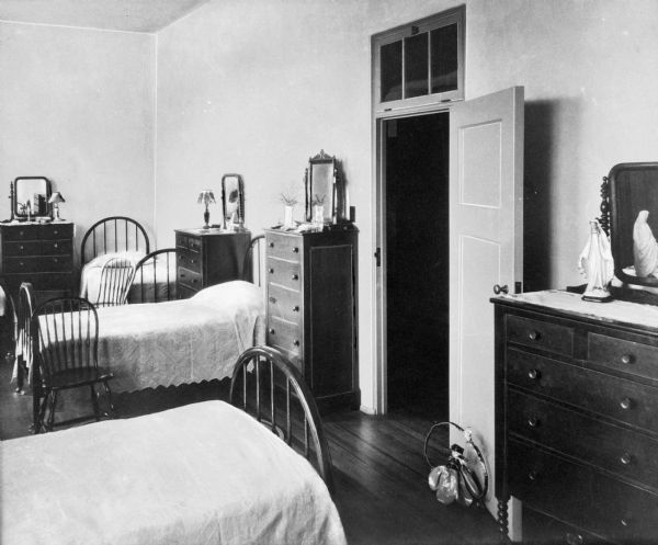 A view of a dormitory at Sacred Heart Villa, with many stations for students consisting of a bed and dresser.  Each dresser has different objects on them, including flowers and a statue of Mary.