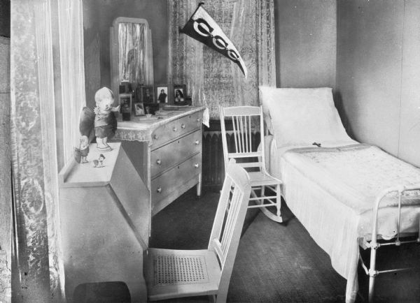 A view of a bedroom with bed, dresser, and desk.  The windows have lacy curtains and the top of the desk contains multiple Kewpie dolls
