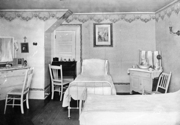 A view of a two-person bedroom at the Academy of Jesus and Mary.  The room is furnished with two beds, a desk, dresser, and wash stand.