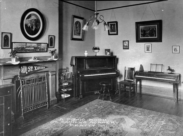 A view of the piano room at St. Xavier's Academy. An upright piano and a smaller keyboard stand next to each other along a wall decorated with framed pictures. A pennant reading "St. Xavier" hangs over the fireplace, and a carpet lies on the floor. Caption reads: "A Piano Room, St. Xavier's Academy Beatty. PA."