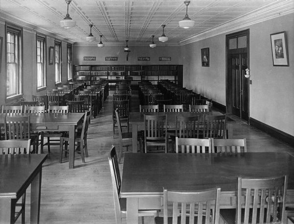 A view of the Study Hall at Putnam Catholic Academy. The room contains both small tables for one or two people and large tables for groups.  Framed artwork hangs on the walls and bookcases are in the background.