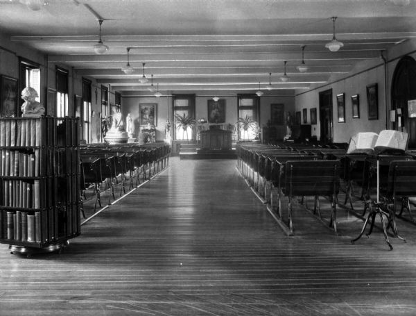 A view of the study hall at St. Joseph's Academy of Maine, which appears to be connected to or double as a chapel. A bookshelf holding a bust sculpture is standing in the foreground. Chairs are lined up behind it, facing an altar with a portrait of Jesus hanging on the wall above it.
