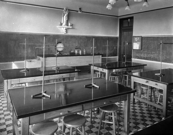 Slightly elevated view of a science classroom at Putnam Catholic Academy. Two chalkboards line the walls and stools stand at laboratory tables throughout the room. A table with an attached sink at the front of the class is set with various instruments and the room is overlooked by a statue of the Virgin Mary.