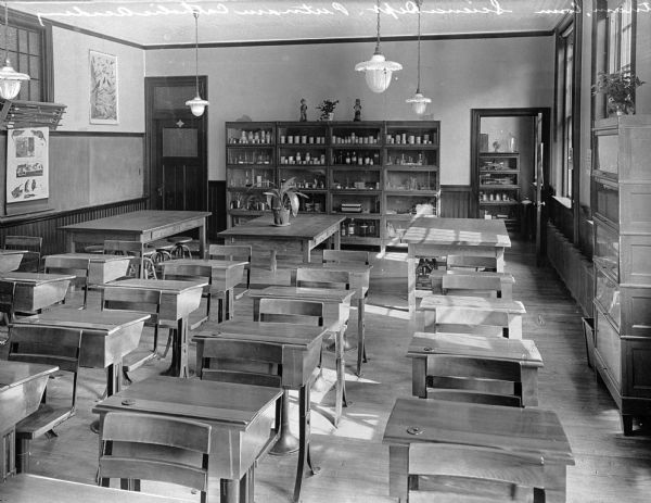 Slightly elevated view of a science room at Putnam Catholic Academy, looking out over the desks toward the back of the classroom. In the back is a large glass-door cabinet that contains various bottles and instruments. Three large work tables sit near the cabinet, the middle one holding a potted plant.
