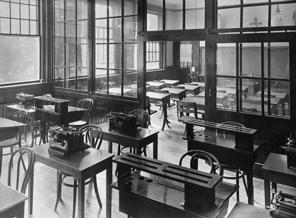 View across desks with typewriters in a classroom. A windowed partition separates it from a classroom with desks facing a blackboard on the other side.