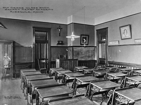 Rows of desks lined up inside an eighth grade classroom at the Sacred Heart Academy. A teacher's desk is by the front chalkboard, standing beneath a crucifix hanging on the wall. Framed artwork and a Sacred Heart pendant decorate the walls. Caption reads: "8th Grade Class Room, Sacred Heart Academy Missoula, Mont."