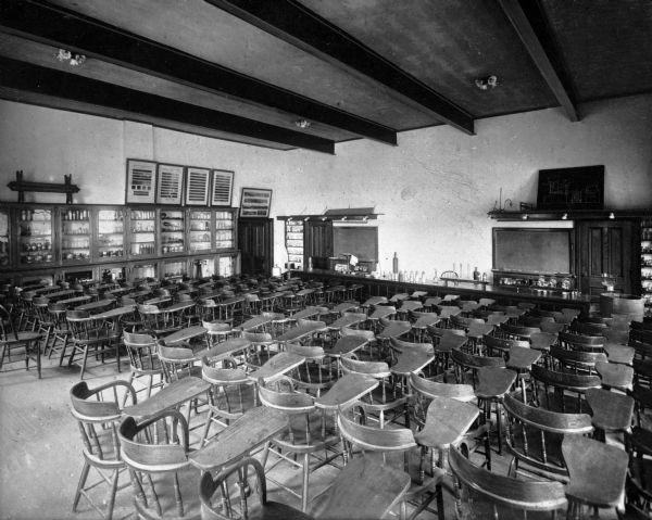 A view of a classroom at Lafayette College looking toward the front of the room. A long demonstration table with bottles takes up most of the front, and rows of chairs fill the rest of the space. To the left is a long cabinet with glass doors that contains books and scientific  instruments. Framed prints sit on top of the cabinet.