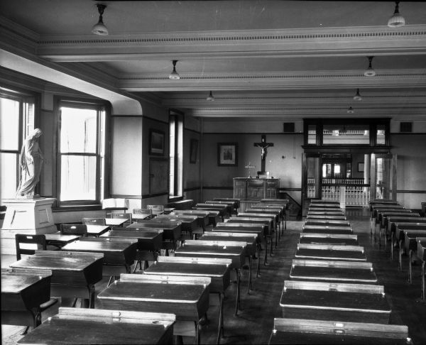 A view of the study room at St. Joseph's college, lined with rows of desks. This room appears to double as a worship space, as the desk at the front of the room is elevated and a small crucifix sits on its surface. A large wall-mounted crucifix is behind it. A statue of Mary stands in the window alcove.