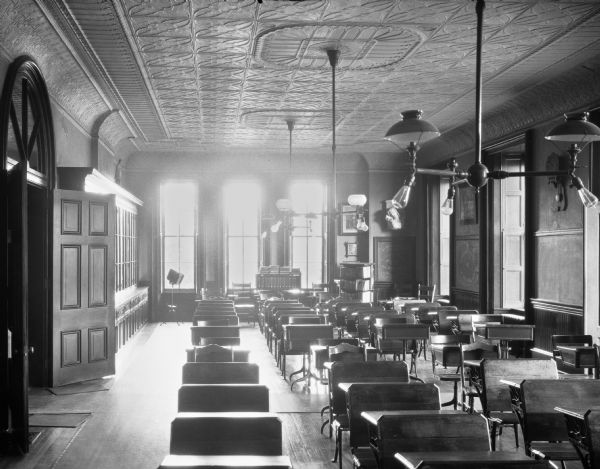 A view looking over rows of desks in the novitiate classroom at Mount St. Joseph. Light fixtures hang from the tin ceiling and a double door entrance is located along the left wall. A long storage cabinet stands next to the doors and bookcases are near the windows.