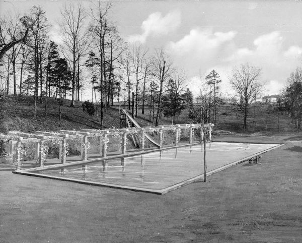 A view of a swimming pool beside a hill at Bessie Tift College. Next to the pool is a vine-covered pergola and a slide. College buildings are visible in the distance.