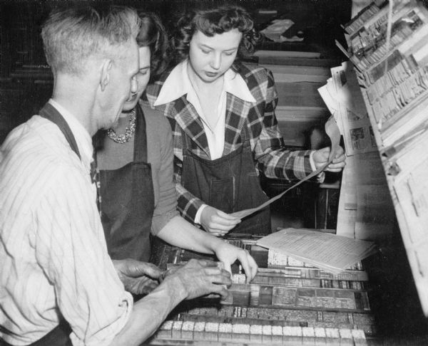 Two female students stand with a male teacher during a Practical Journalism Training class in the newspaper shop at Westminster College. The students and teacher all wear aprons while typesetting a newspaper page.