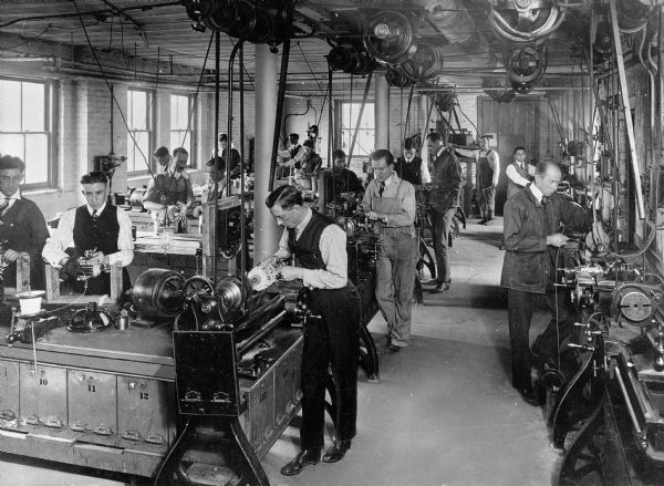 Students stand at work stations in the machine shop at Bliss Electrical School. They men all appear to be wiring something on a mount.
