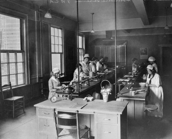 Students sitting and standing at workstations set at long tables during a cooking class at the Young Women's Christian Association. A teacher's desk in the foreground contains a basket of flowers and a skillet.