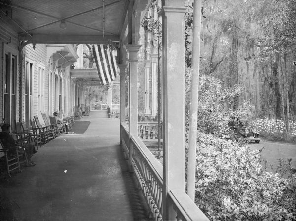 People sit in rocking chairs on the porch of the Pine Forest Inn. Flowering bushes and shrubs surround the porch and an automobile is driving along the road to the right.