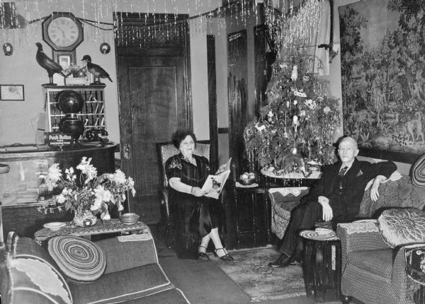 A view of the Ideal Hotel lobby during the Christmas season. A small, decorated tree is on a table near the front desk, and a man and women sit on a chair and sofa on either side.
