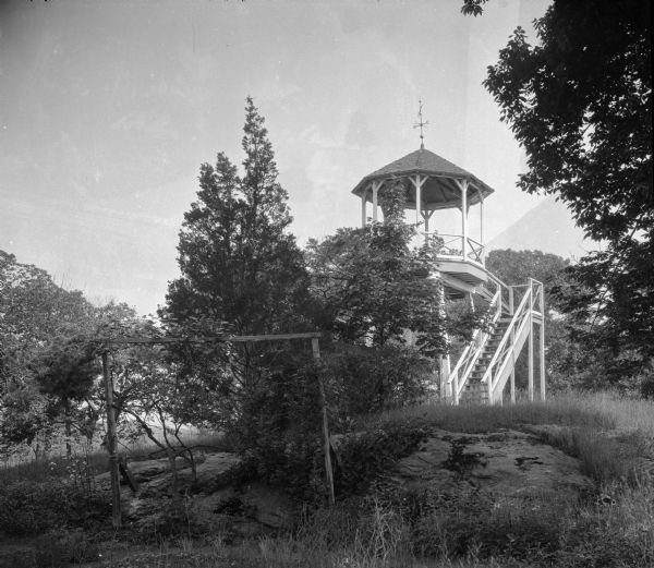 A view of the open-air pavilion at Sheldon House Hotel. The pavilion stands in a wooded area with mountains visible in the distance. A set of wooden stairs leads up to the structure's platform, and the roof has a weather vane at its peak.